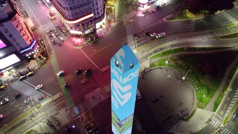 Aerial-orbit-of-the-Obelisk-in-Buenos-Aires,-with-the-colors-of-the-Argentine-flag,-cheering-the-national-soccer-team-in-the-Qatar-2022-World-Cup,-Corrientes-Avenue-and-neon-lights-advertisements