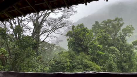 Starke-Regenfälle-Vom-Hotel-Lodge-Balkon-Mit-Tropischer-Vegetation-Während-Des-Monsuns-Mit-Berg-Im-Hintergrund,-Der-Durch-Den-Nebel-Gesehen-Wird