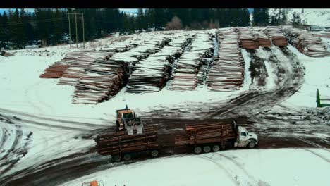Wheel-loader-with-grapple-bucket-unloading-log-truck-and-maneuvering-through-large-log-piles-at-Canadian-sawmill-yard-in-winter