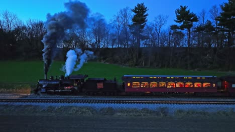 A-Drone-Parallel-Night-View-of-a-Steam-Passenger-Train-Stopped-in-Farmlands-Blowing-Lots-of-Smoke-Seeing-the-Lights-in-the-Coaches