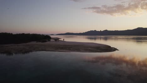Goldener-Sonnenuntergang-Aus-Der-Luft-über-Den-Hügeln-Der-Komodo-Inseln-Mit-Silhouettierten-Touristen,-Die-In-Der-Abenddämmerung-An-Einem-Strand-Spazieren-Gehen