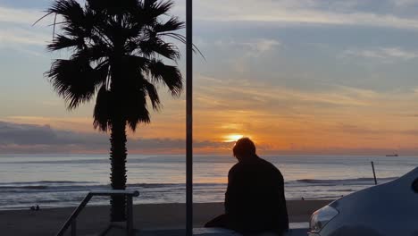 Silhouette-of-a-man-smoking-against-an-orange-sunset,-a-young-man-smokes-a-cigarette-with-the-beach-and-waves-behind