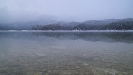 Snowy-misty-ethereal-woodland-reflections-on-lake-Eibsee-shoreline,-Bavaria
