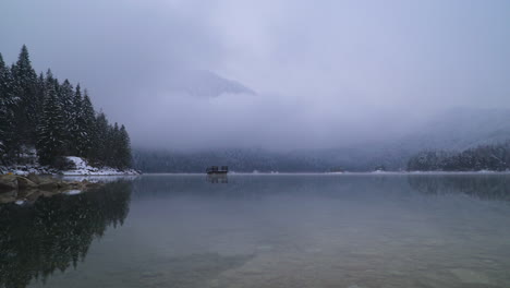 Snowy-woodland-trees-reflections-in-lake-Eibsee-misty-ethereal-mountain-shoreline