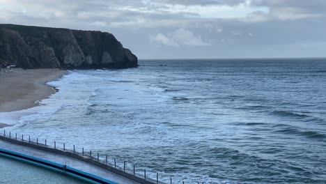 wide-view-Of-Beautiful-Cliff-And-Turquoise-Sea-On-A-cloudy-Day-with-some-surfers-enjoying-small-waves