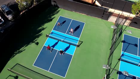 Pickleball-match-on-blue-outside-court,-foursome-playing-the-sport,-drone-rising-over-players
