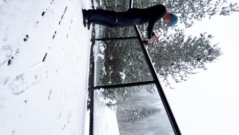 Vertical-video-of-white-male-in-snow-gear-making-coffee-on-porch-covered-in-snow-in-a-pinewood-forest
