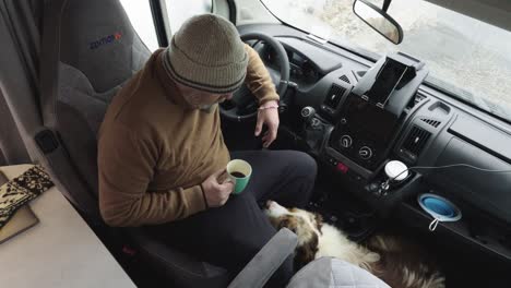 Older-man-drinking-coffee-or-tea-in-campervan,-dog-inside,-top-down-view