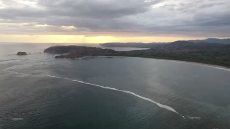 Coastline-of-Costa-Rica-from-the-Pacific-Ocean-at-sunset-with-rocky-peninsula-and-shallow-reef,-Aerial-flyover-shot