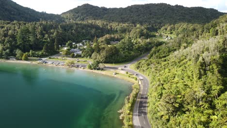 Lakeside-holiday-campground,-blue-lake,-palm-forest-road-and-tourist-on-paddle-bord-4k-drone-shot-in-New-Zealand