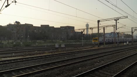 Side-view-of-railway-yard-from-a-moving-train-slowly-leaving-junction,-local-train-standing-and-waiting-for-signal-on-other-line-with-old-rustic-buildings-at-background
