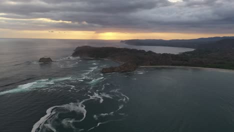 Water-Maelstrom-forming-in-the-coastline-of-Costa-Rica-in-Pacific-Ocean-at-sunset-with-rocky-peninsula,-Aerial-flyover-shot
