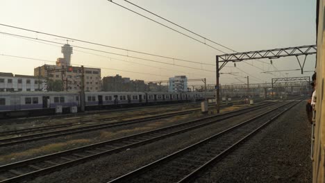 Side-view-of-railway-yard-from-a-moving-train-slowly-leaving-junction,-local-train-standing-and-waiting-for-signal-on-other-line-with-old-rustic-buildings-at-background
