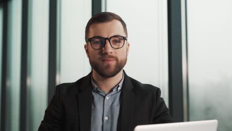 Portrait-of-a-smiling-young-man-wearing-glasses-on-the-background-of-an-office-with-panoramic-windows