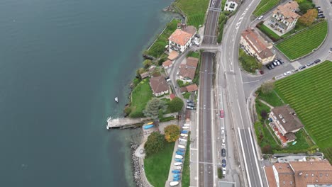 aerial-tilt-up:-road-and-houses-next-to-geneva-lake-and-turquoise-water,-Switzerland
