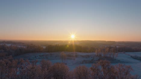 Aerial-establishing-view-of-a-rural-landscape-in-winter,-snow-covered-countryside-fields,-cold-freezing-weather,-sunset-with-golden-hour-light,-wide-drone-shot-moving-backward-low-ovwe-trees