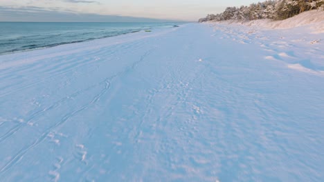 Aerial-footage-of-beach-covered-with-snow,-sunny-winter-day-before-the-sunset,-golden-hour,-Nordic-woodland-pine-tree-forest,-Baltic-sea-coast,-wide-drone-shot-moving-forward-low