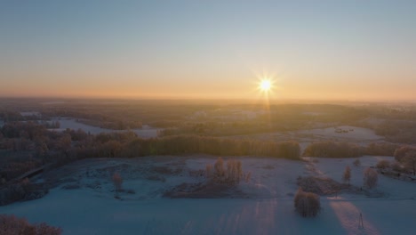 Aerial-establishing-view-of-a-rural-landscape-in-winter,-snow-covered-countryside-fields,-cold-freezing-weather,-sunset-with-golden-hour-light,-wide-drone-shot-moving-backward