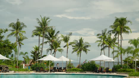 Hotel-Guests-On-The-Poolside-With-Loungers,-Umbrella,-And-Palm-Trees