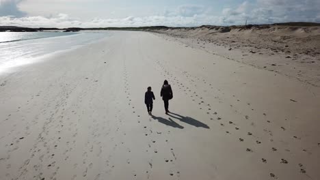 aerial-push-in:-two-people-walk-on-a-large-sandy-beach-in-Connemara,-Ireland