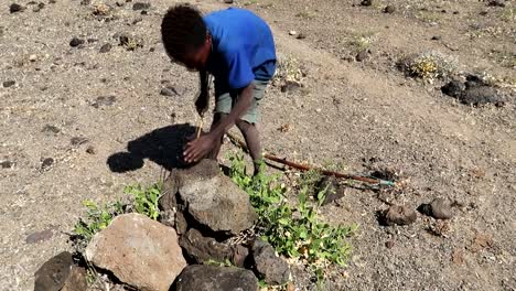 Local-African-boy-sharpening-a-self-made-arrowhead-against-a-rock-in-the-Tanzanian-savanna