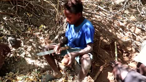 Full-shot-of-Hadzabe-tribe-boy-peeling-Boabab-tree-root-sitting-on-ground