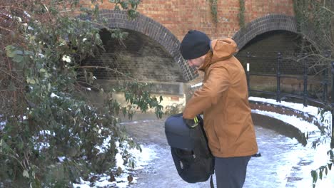 Warm-dressed-male-putting-drone-carry-bag-into-rucksack-beside-snow-Brooklands-lake-archway