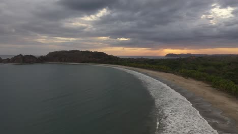 Curved-half-moon-coastline-of-Costa-Rica-with-soft-waves-on-sandy-shore-at-sunset,-Aerial-flyover-shot