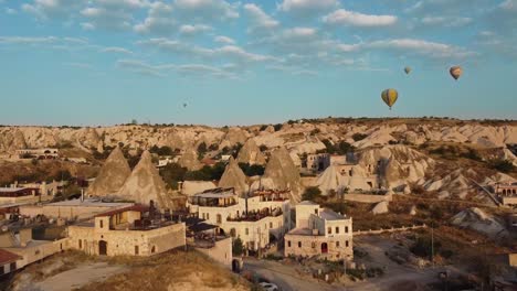 Flight-over-the-houses-of-Goreme,-with-hot-air-balloons-in-the-sky,-in-Cappadocia-of-Turkey