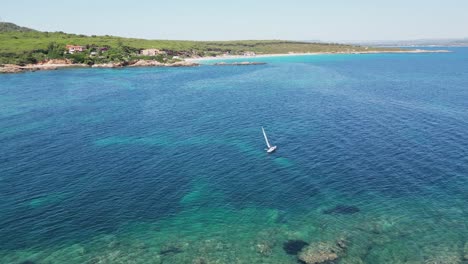 Solo-Sailor-sails-along-Cala-Spinosa-Beach-in-Sardinia,-Italy---4k-Aerial-Backwards