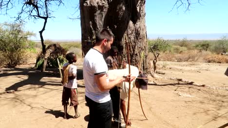 Touristenmann,-Der-Versucht-Zu-Lernen,-Wie-Man-Einen-Bogen-Benutzt,-Um-Mit-Dem-Hadzabe-Stamm-In-Der-Afrikanischen-Savanne-Zu-Jagen