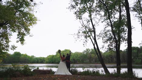 Unrecognizable-newly-wed-couple-raising-arms-up,-celebrating-marriage-outdoors-in-forest-by-river