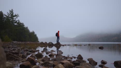 Man-in-red-rain-jacket-stands-on-rocks-near-a-lake-with-fog-and-mountains-in-Acadia-Park-in-Maine
