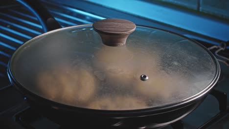 Hand-covering-fritters-in-a-black-frying-pan-with-a-glass-transparent-lid-on-a-stove