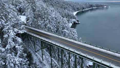 Orbiting-aerial-shot-of-an-empty-bridge-over-the-ocean-connecting-to-land-covered-in-fresh-snowfall