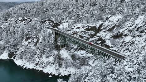 Drone-shot-of-a-red-car-driving-across-an-elevated-bridge-with-snow-everywhere