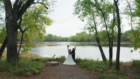 Aerial,-newly-wed-heterosexual-couple-raising-arms-up-celebrating-marriage-in-outdoor-forest-by-a-river