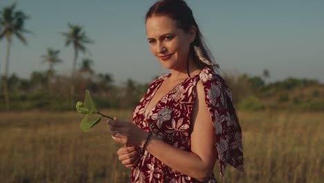 Beautiful-woman-in-a-floral-print-dress-standing-in-a-field-holding-a-leaf-in-her-hands-and-smiling-at-the-camera