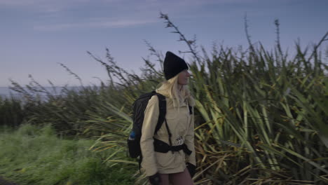 Attractive-blond-female-traveler-walking-on-path-between-tall-grass,-Waipapa-Point
