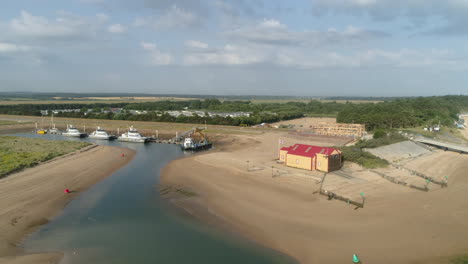 Pullback-Drone-Shot-of-Wells-Next-The-Sea-Creek-at-Low-Tide-with-Old-Lifeboat-House-and-Working-Windmill-Maintenance-Boats-in-North-Norfolk-UK-East-Coast