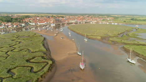 Establishing-Drone-Shot-Coming-in-towards-Wells-Next-The-Sea-Coastal-Town-with-Salt-Marsh-and-Creek-and-Sailing-Boats-at-Low-Tide-in-North-Norfolk-UK-East-Coast