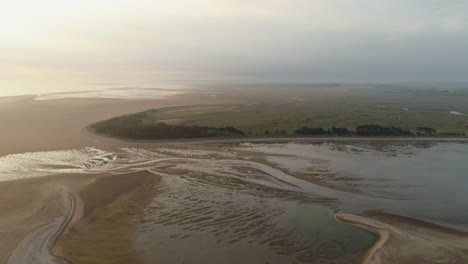 Cinematic-Establishing-Drone-Shot-at-Low-Tide-Towards-Sandy-Beach-with-Trees-and-Salt-Marsh-Behind-at-Sunrise-in-North-Norfolk-UK-East-Coast