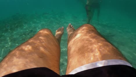 Underwater-subjective-perspective-of-man-legs-floating-in-clear-transparent-sea-water-beneath-surface-with-people-in-background