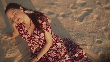 Woman-sitting-in-the-sad-at-the-beach-in-a-dress-with-eyes-closed-enjoying-the-sunlight