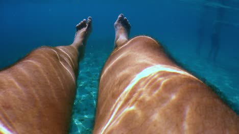 Underwater-personal-perspective-view-of-man-legs-floating-in-clear-transparent-sea-water-beneath-surface-with-blue-background