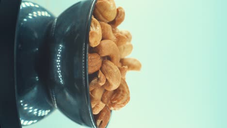 A-vertical-close-up-shot-of-a-roasted-salty-cashews-in-a-black-shiny-bawl-on-a-rotating-stand,-slow-motion,-4k,-man-hand-taking-a-cashew-of-the-bawl