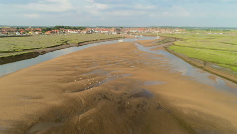 Drone-Shot-Down-Creek-at-Low-Tide-with-Birds-Flying-and-Wells-Next-The-Sea-Coastal-Town-in-Background-in-North-Norfolk-UK-East-Coast
