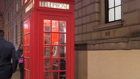 Red-Telephone-Booth-On-The-Sidewalk-With-People-Walking-At-Night-In-London,-UK