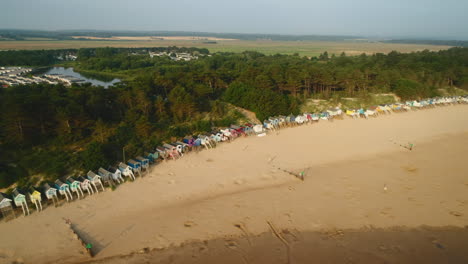 Drone-Shot-Along-Sandy-Beach-with-Beach-Huts-and-Woodland-Behind-on-Sunny-Morning-in-Wells-Next-The-Sea-North-Norfolk-UK-East-Coast