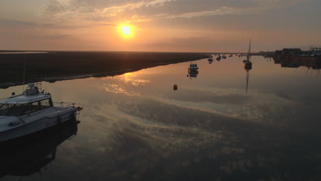 Low-Drone-Shot-Over-Still-Water-with-Small-Boats-and-Sailing-Boats-on-Creek-at-High-Tide-with-Salt-Marsh-and-Stunning-Sunrise-in-North-Norfolk-UK-East-Coast